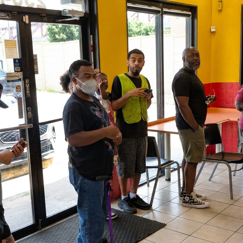 A group of people stands inside a brightly colored restaurant near the entrance, windows, tables, and chairs.