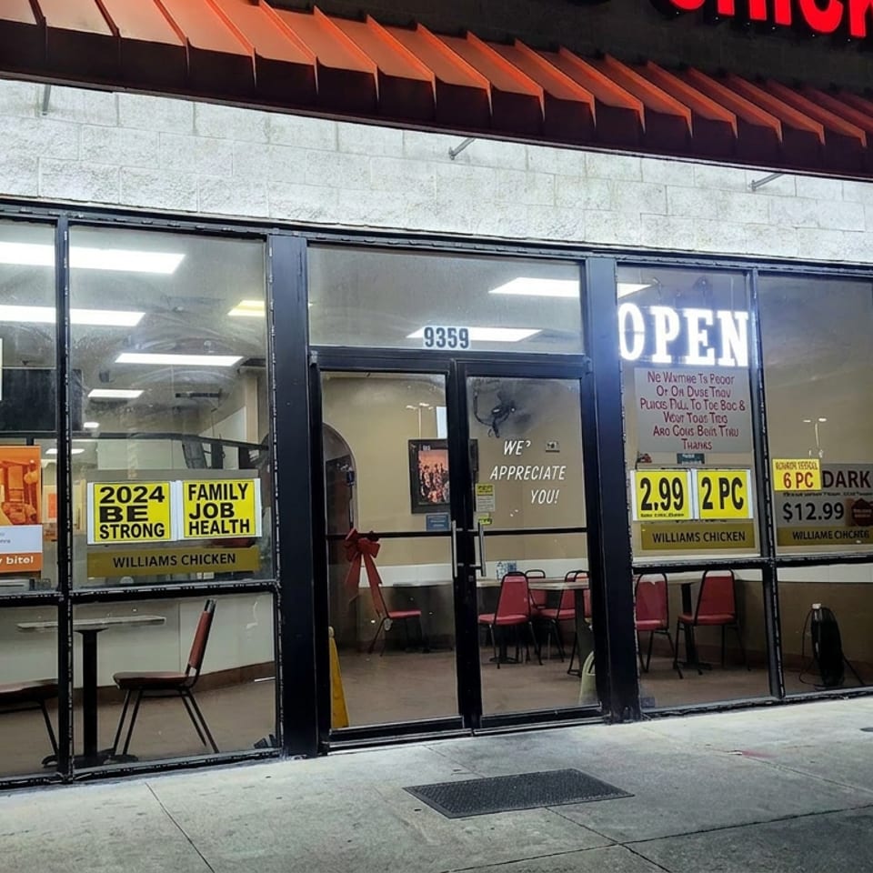 Exterior of a Williams Chicken restaurant with illuminated open sign, promotional window posters, and visible seating inside.