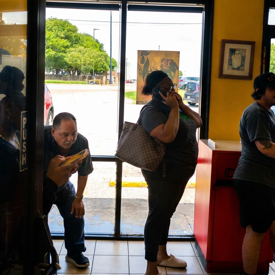 Restaurant entrance with several patrons waiting near the glass doors, including a woman on the phone and a man checking his phone.
